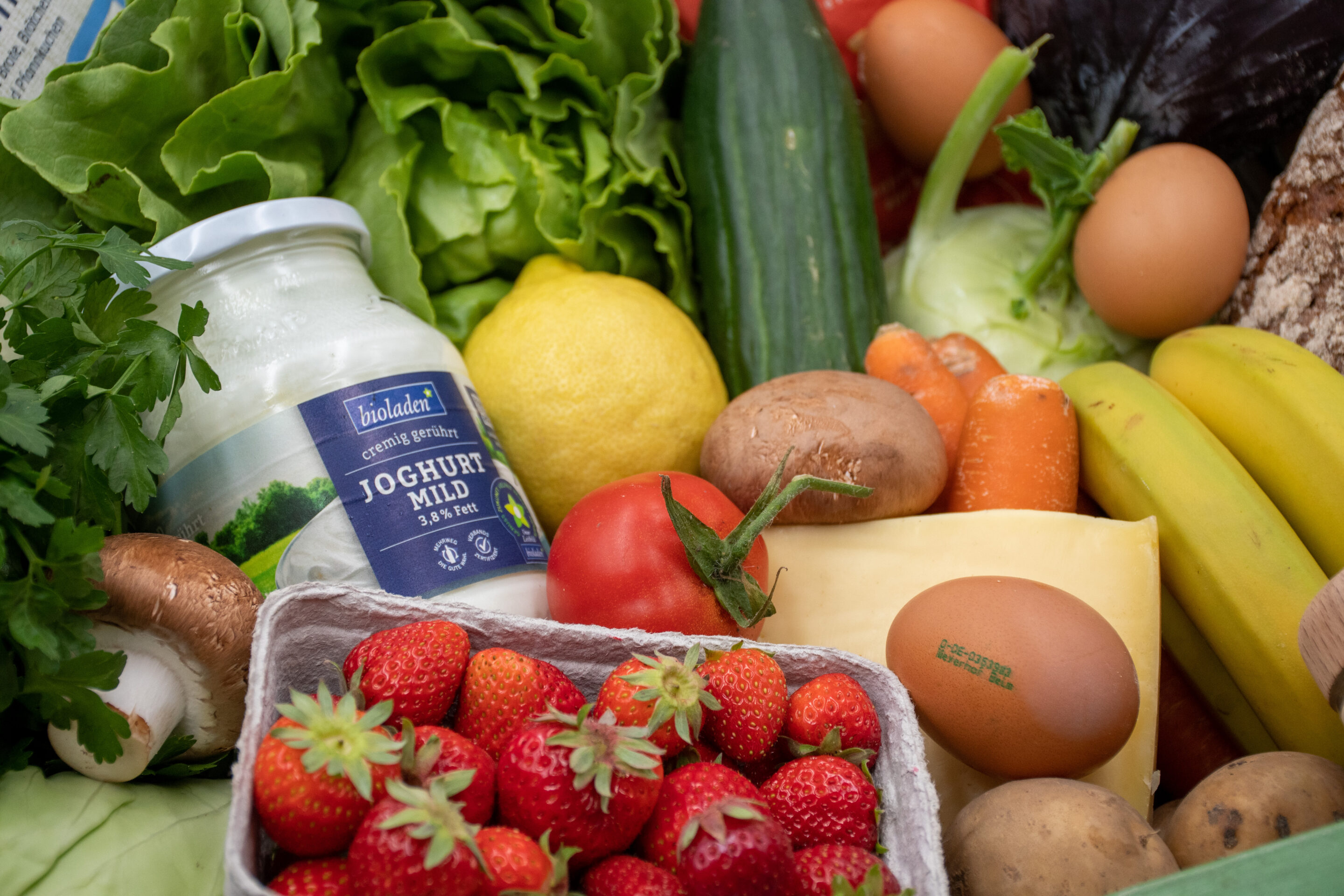 Verschiedene frische Lebensmittel wie Erdbeeren, Tomate, Zitrone, Gurke, Karotten, Salat, Bananen, Kartoffeln, Eier, Käse und ein Glas Joghurt