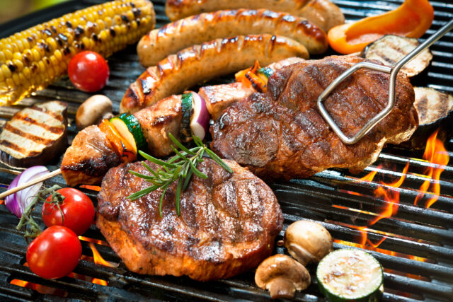 Gegrilltes Steak mit Rosmarin, Würstchen, Maiskolben, Gemüse und Pilzen auf Grillrost über offenem Feuer