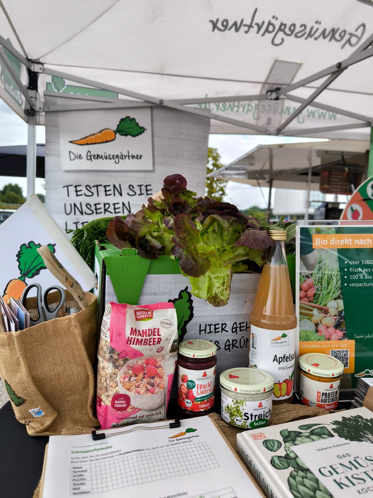 Marktstand mit frischem Salat in grünem Karton, Apfelsaftflasche, Mandel-Himbeer-Müsli, Marmelade, Brotaufstrich und Informationsflyern unter einem Zelt mit Aufschrift 'Die Gemüsegärtnerei'