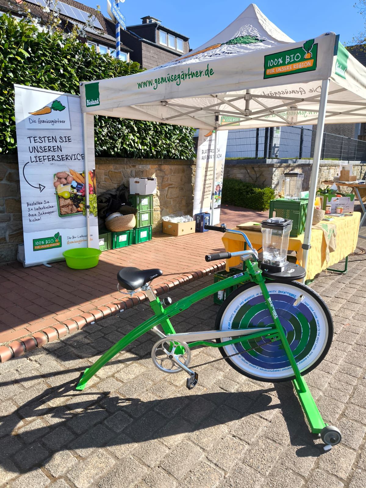 Grünes Smoothiebike mit großem Rad und Pedalen vor einem weißen Zelt mit der Aufschrift 'www.gemüsegärtner.de' und '100% BIO' auf einem Pflastersteinweg