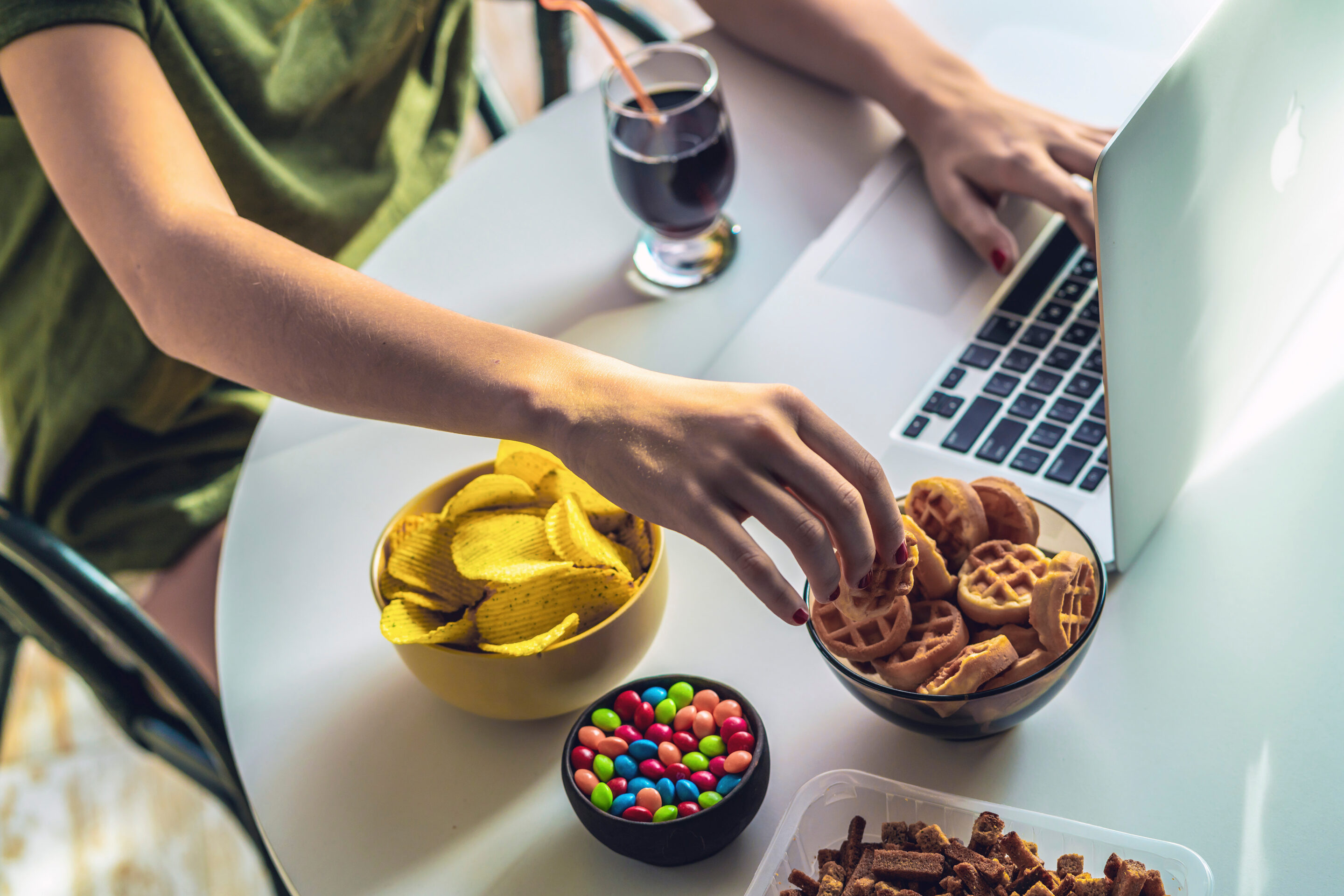 Mädchen arbeitet am Computer und isst Fast Food. Ungesunde Lebensmittel: Chips, Cracker, Süßigkeiten, Waffeln, Cola.