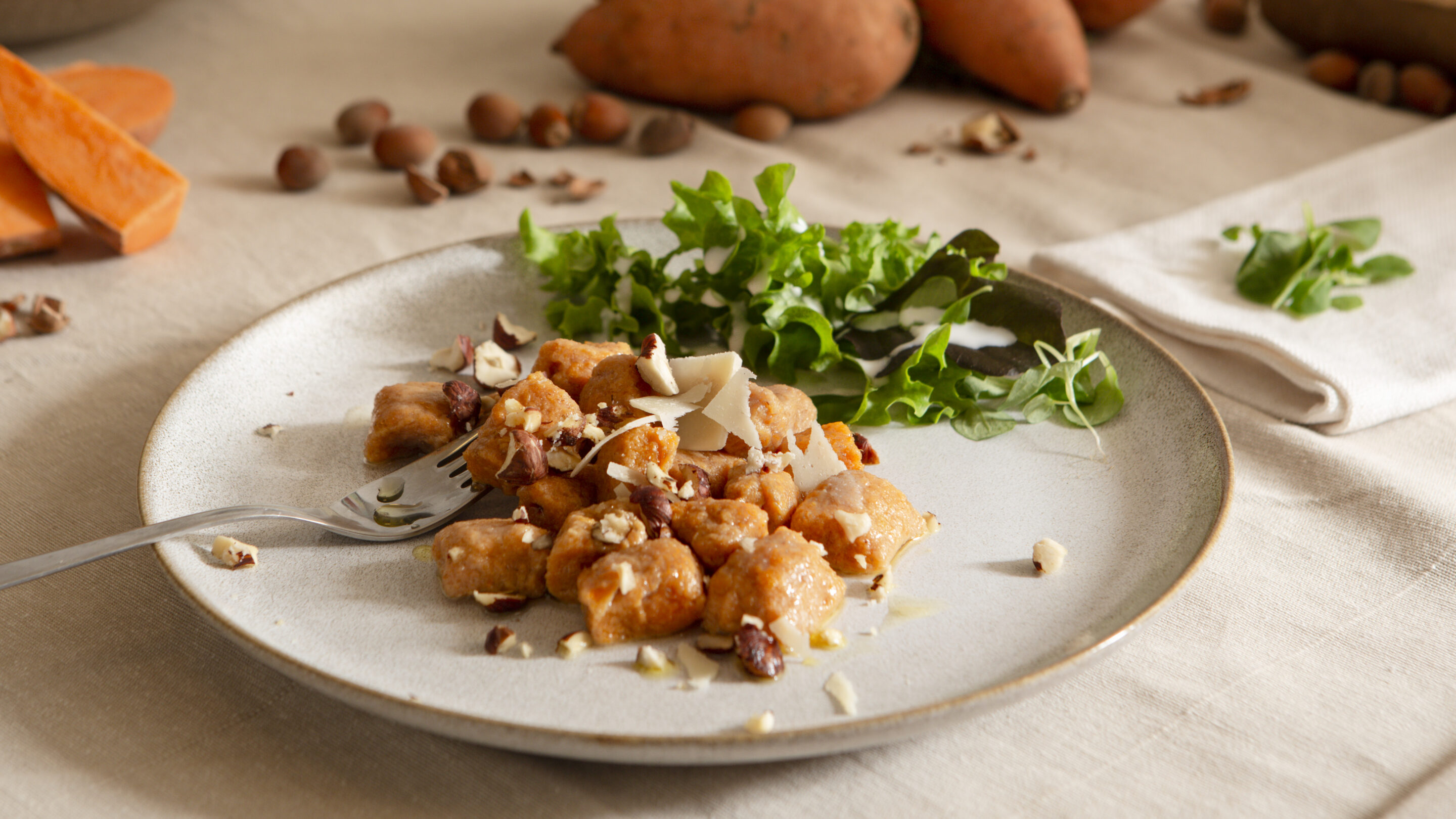 Gekochte Gnocchi mit gehackten Nüssen und grünen Blättern auf einem hellen Teller, daneben eine Gabel und ein weißes Stofftuch.