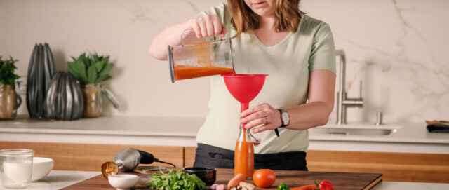 Frau gießt mit Trichter eine orangefarbene Flüssigkeit aus einem Mixbehälter in eine Flasche auf einer Küchenarbeitsplatte.