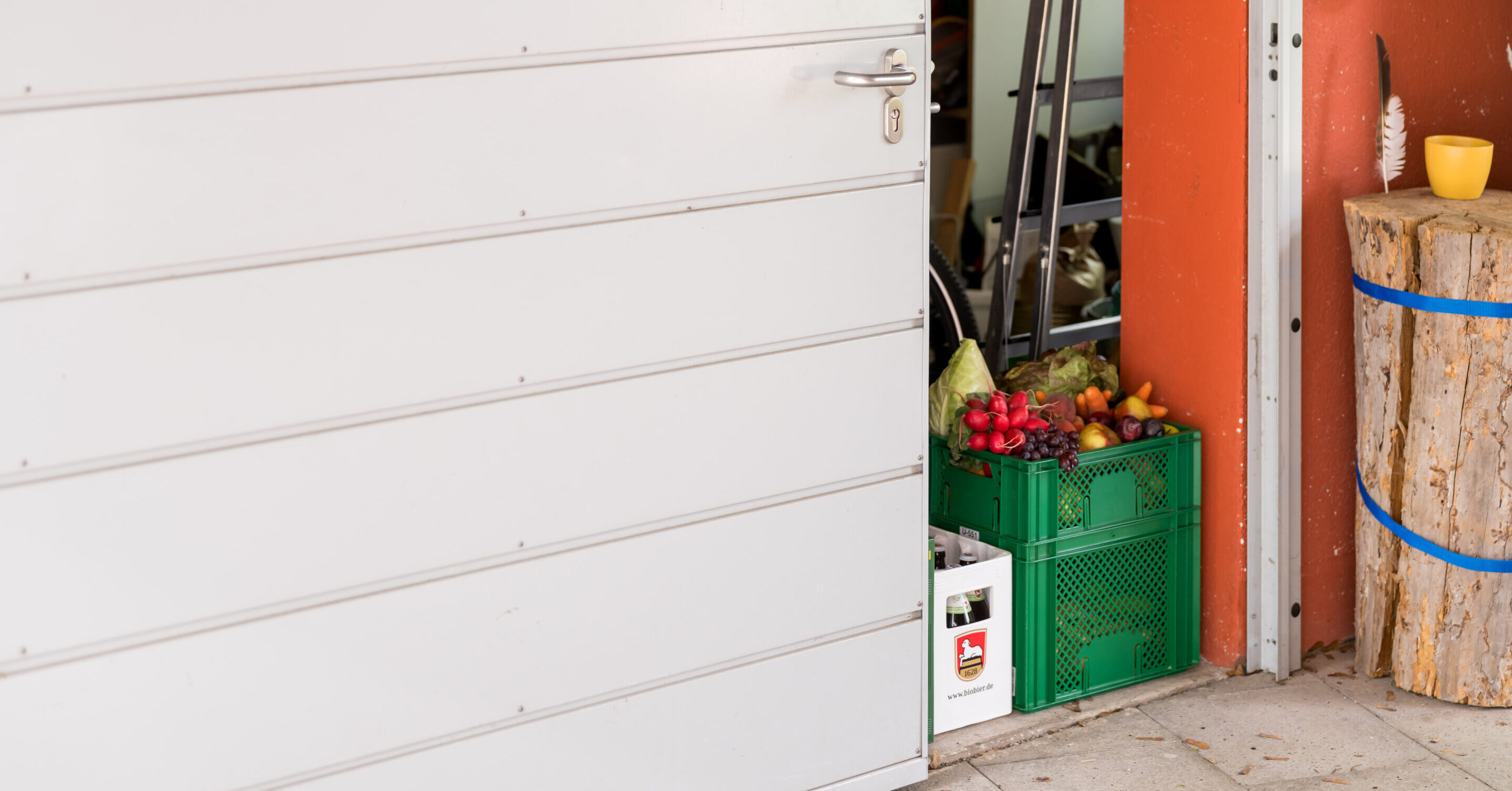 Eine Tür, welche leicht offen steht und da stehen zwei Kisten mit Lebensmitteln übereinander und eine Kiste Bier daneben.