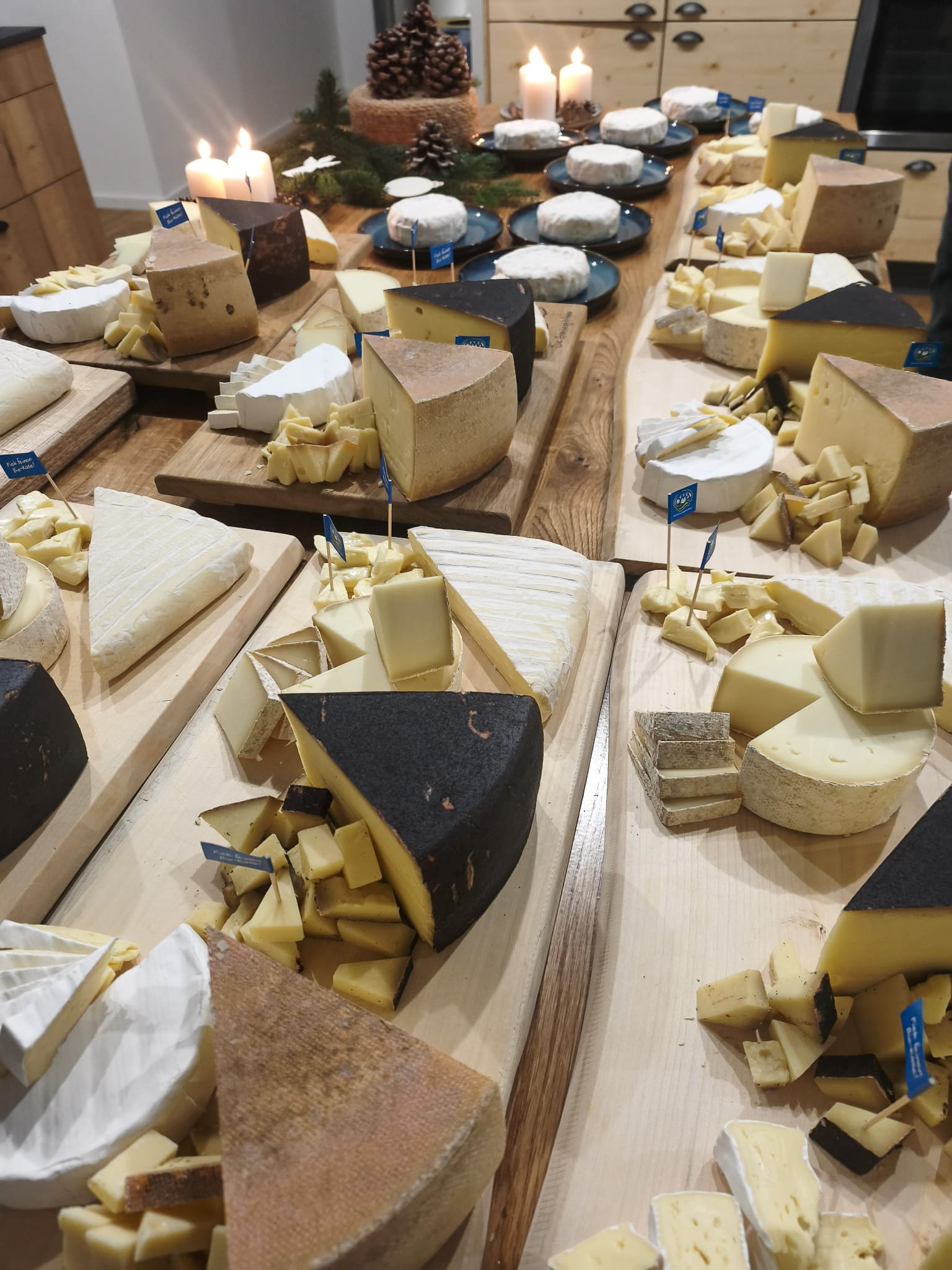 Verschiedene Käsesorten in Stücken und Scheiben auf Holzplatten angerichtet, im Hintergrund brennende Kerzen und Tannenzapfen.