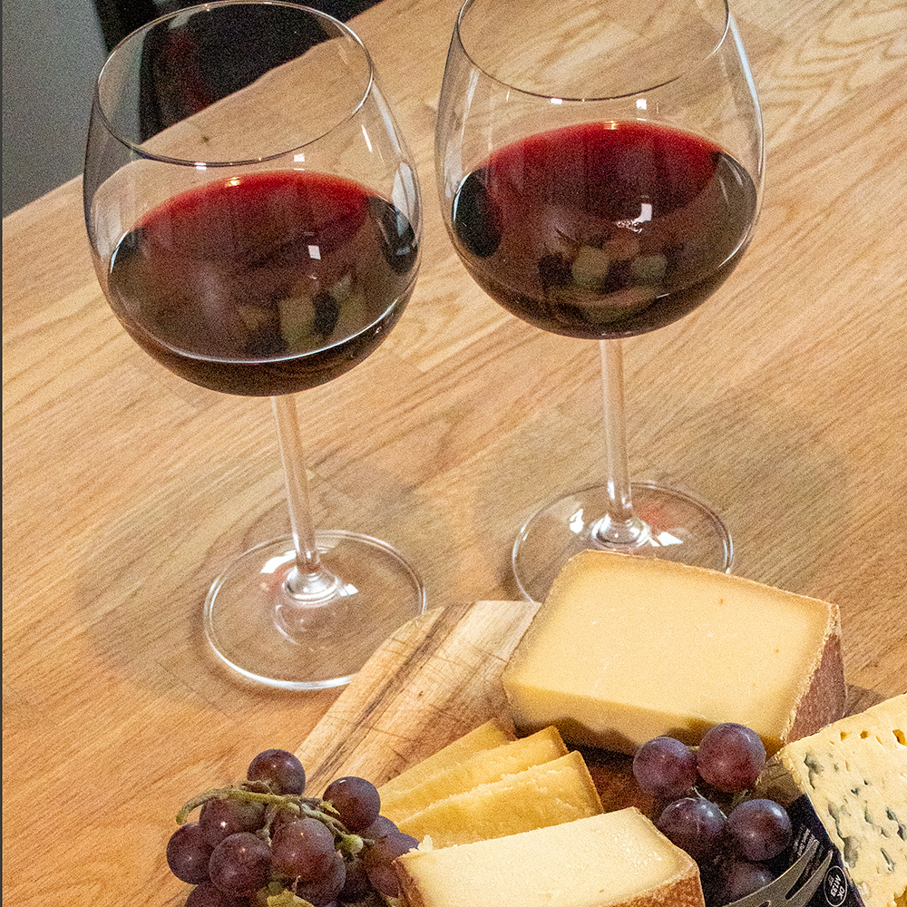 Zwei Weingläser mit Rotwein, verschiedene Käsesorten und Trauben auf einem Holzbrett auf einem Holztisch.